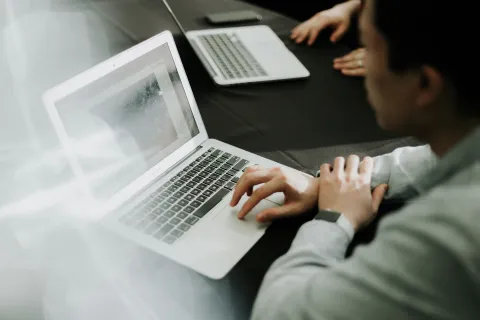 Person working on a laptop at a table with another laptop and person in the background, focusing on the screen.