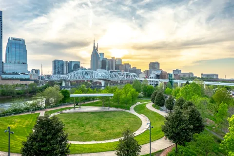 Nashville skyline at sunset with green park, river, and John Seigenthaler Pedestrian Bridge in view