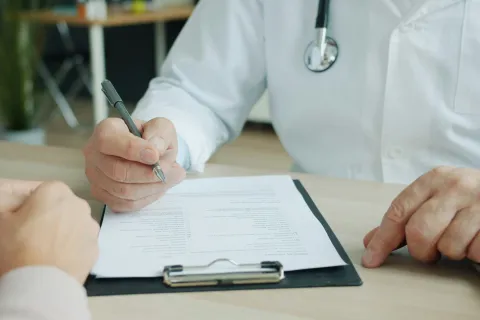 Doctor in white coat with stethoscope writing on medical form during patient consultation.