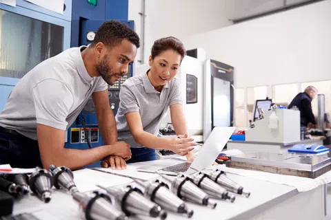 Two engineers collaborate on a laptop in a manufacturing workshop with machinery and metal parts on the table.