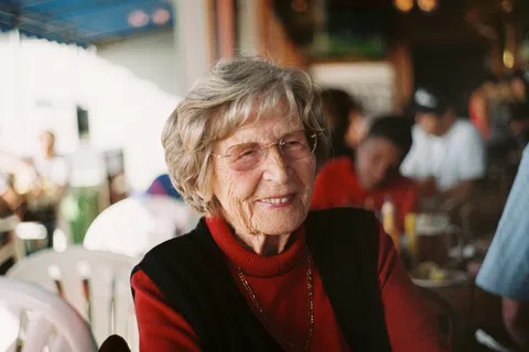 Smiling elderly woman wearing glasses and a red turtleneck sweater sitting in a busy indoor setting.