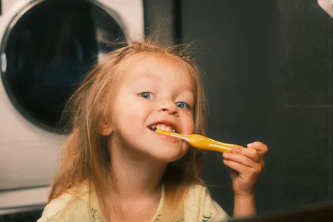 Young child brushing teeth in bathroom with washing machine in the background looking at mirror.