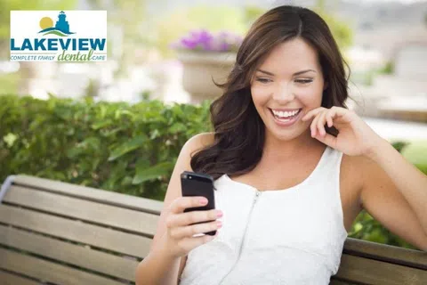 Happy young woman smiling while looking at her smartphone outdoors on a park bench with greenery background