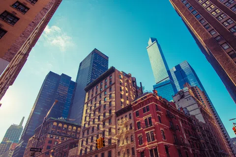 View looking up at a city street with diverse tall buildings under a clear blue sky in bright sunlight