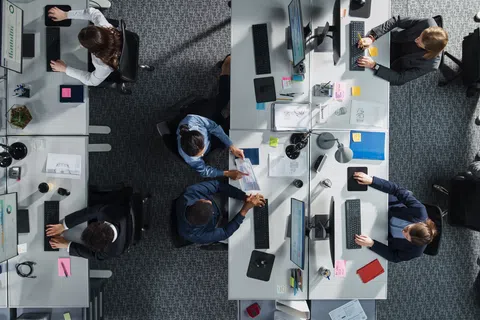 Overhead view of professionals collaborating and working at desks with computers and documents in modern office space.