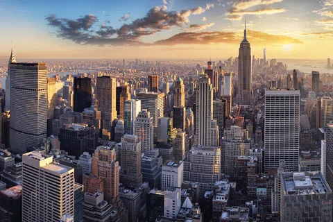 New York City skyline with Empire State Building and One World Trade Center at sunset under partly cloudy sky.