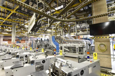 Modern industrial printing machines connected by overhead conveyor belts inside a spacious factory setting