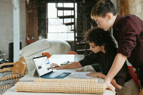 Two designers collaborating at a table using a laptop and reviewing architectural plans in a modern studio.