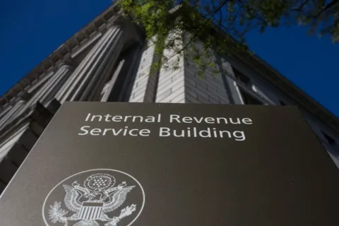 Sign for the Internal Revenue Service Building with part of the government building and tree against blue sky.