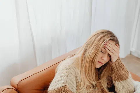 Woman with blonde hair sitting on a brown couch holding her forehead, appearing stressed or tired in a bright room.