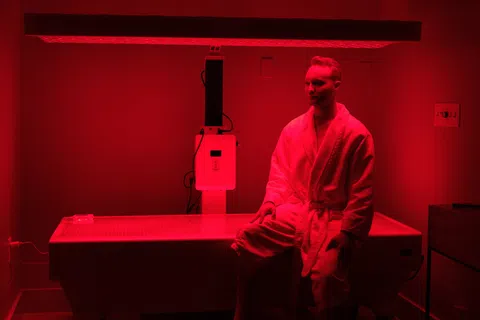 Man in white robe sitting on a bed under red light therapy device in a dim room