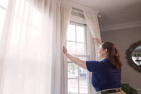 Woman opening white curtains to let in natural light through a large window in a cozy room.