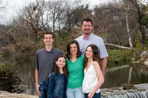 Smiling family of five posing outdoors by a small waterfall and bare trees in a natural setting