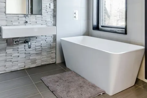Modern bathroom with freestanding white tub, wall-mounted sink, textured tile wall, and window blinds.