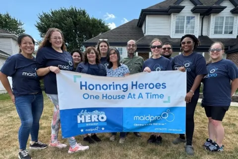 Group of people holding a banner reading Honoring Heroes One House At A Time outside a suburban house on a sunny day