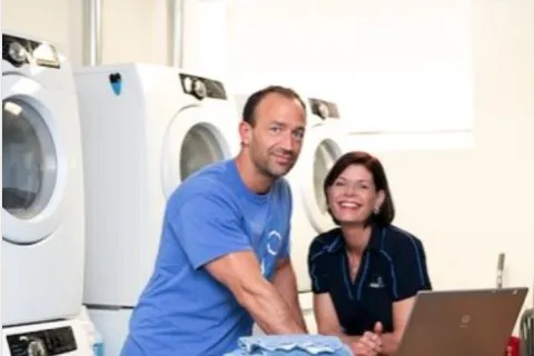Two people in a laundry room folding clothes near stacked washers and dryers with cleaning supplies nearby.
