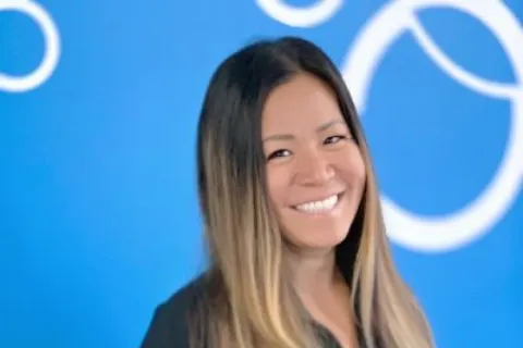 Smiling woman with long ombre hair wearing a black top standing against a blue wall with white circular patterns.