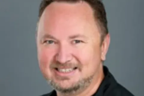 Portrait of a smiling man with short curly hair and a trimmed beard wearing a black zip-up jacket.