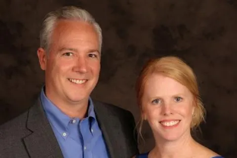 Smiling man in gray suit and blue shirt with woman in sleeveless blue dress against dark background