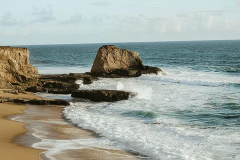 Rocky coastline with sandy beach and waves crashing under cloudy sky at serene ocean shore
