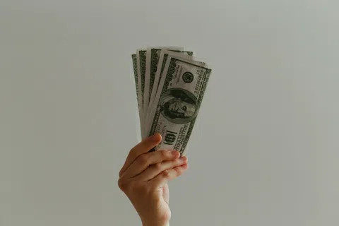A hand holding multiple hundred-dollar bills against a neutral background.