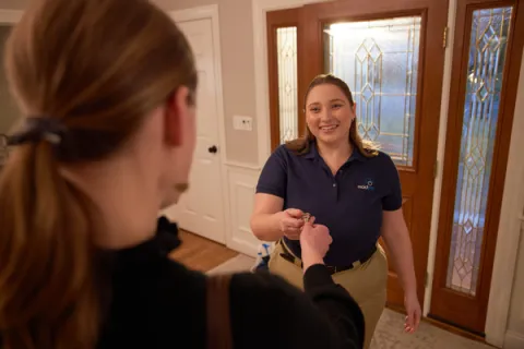 Home service worker handing keys to a smiling customer inside a warmly lit entryway with decorative glass doors.