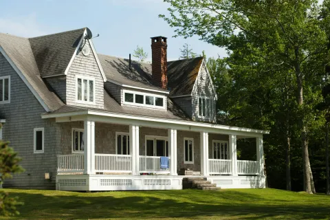 Gray shingle house with white porch, chimney, and green lawn surrounded by trees on a sunny day