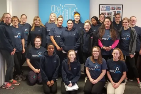Group photo of MaidPro cleaning service team wearing branded shirts in office with blue wall art.