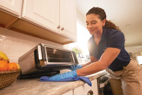 Woman wearing gloves cleaning a kitchen toaster oven on a marble countertop with a blue cloth.