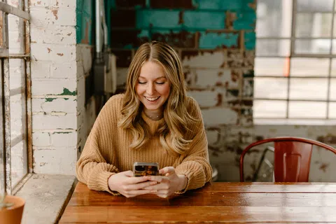 Young woman in a brown sweater smiling while using her smartphone sitting at a wooden table by a window.