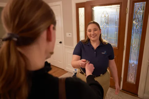 Smiling cleaning service worker handing keys to a woman at a home's front door during a service appointment.