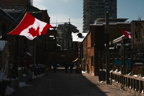 Canadian flags flutter over a narrow cobblestone street lined with brick buildings under a cloudy sky.