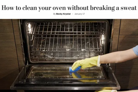 Person wearing yellow gloves cleaning a dirty oven door with a blue sponge in a modern kitchen.