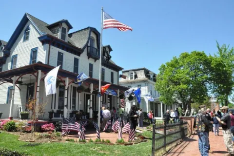 Crowd gathered around historic house decorated with American flags under clear blue sky on a sunny day