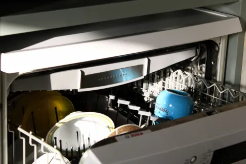 Open dishwasher loaded with plates and a blue mug inside a modern kitchen appliance.
