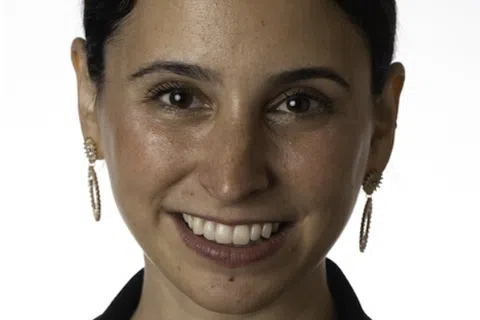 Smiling woman with dark hair in a black outfit wearing earrings and a delicate necklace against a white background