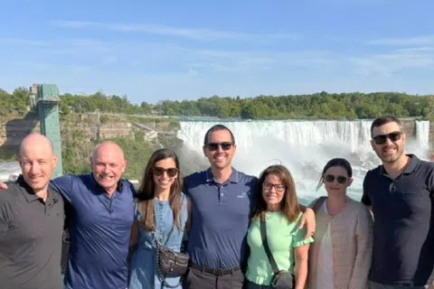 Group of seven people smiling and posing in front of a large waterfall on a sunny day.