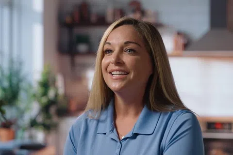 Smiling woman with blonde hair wearing a blue shirt in a bright, blurred kitchen setting.
