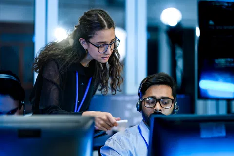 Woman explaining something to male colleague at computer in dimly lit modern office with headsets.