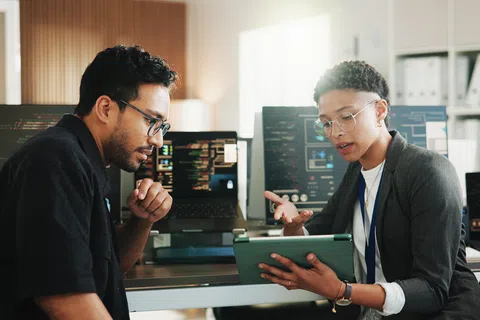 Two diverse coworkers discussing a project using a tablet in a modern office with multiple computer screens.