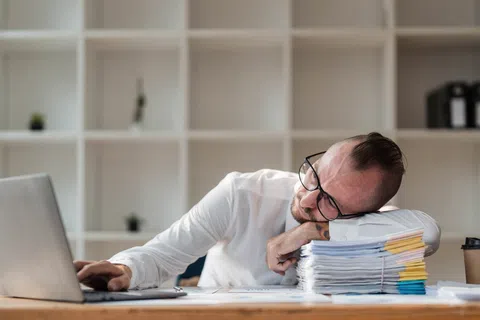 Tired man in glasses resting head on a stack of papers while working on laptop in office.