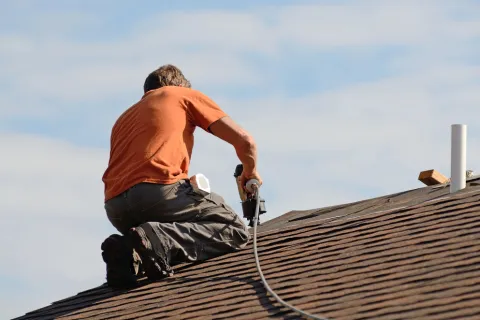 Roofer in orange shirt kneeling on shingled roof using nail gun under a blue sky during roof installation.