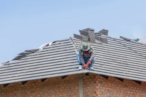 Construction worker installing gray roof tiles on a brick house under a clear blue sky.
