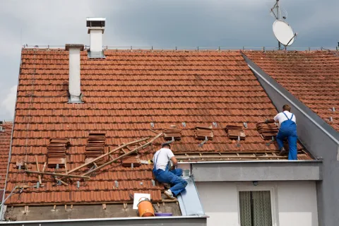 Two workers in blue overalls repairing a red tiled roof on a cloudy day with tools and stacked tiles
