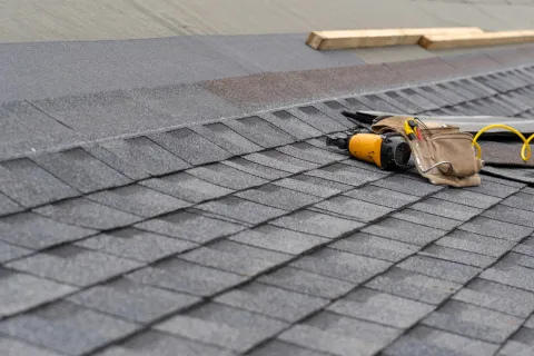 Close-up of newly installed gray asphalt roof shingles with a nail gun and tool pouch on the roof.
