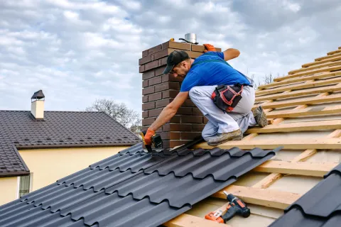 Roofer installing dark metal tiles near brick chimney on house under cloudy sky