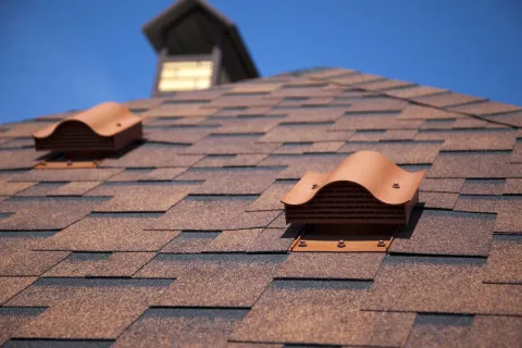 Close-up of a brown shingled roof with two terracotta-colored ventilation caps under a clear blue sky