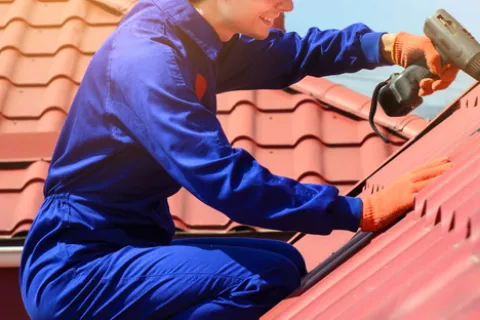 Roofing professional in blue coveralls installing red metal roof panels using a drill on a sunny day