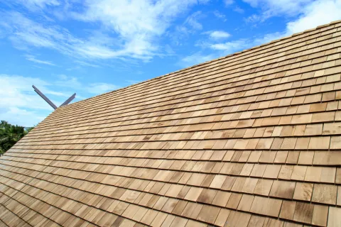 Wooden shingle roof under a partly cloudy blue sky with green foliage in the background.