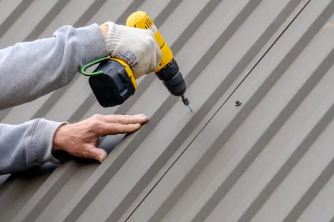 Worker installing screws on a metal roof using a yellow cordless power drill with gloved hand